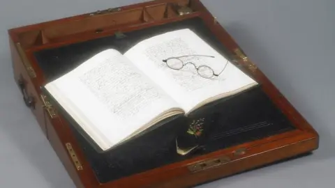 Jane Austen's wooden writing box open with an open book of hand-written text and a pair of spectacles resting on the right hand page