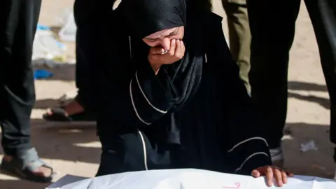 A crying woman wearing dark clothing and a hijab kneels, one hand to her mouth, the other on the shrouded body of a corpse 