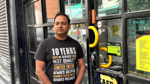 Rajiv Prasad, a man with short, black hair, stands in front of his samosa cafe in King Street Belfast.  He is wearing a black t-shirt with a large logo referring to "10 years in service".  The front of the cafe is constructed of a wall of small windows surrounded by a black wooden frame. 