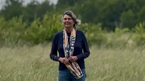 Helen Lewy is pictured standing in a field looking up to the sky. She is an older woman with dark grey hair that is blowing in the wind. Helen is wearing a navy jumper and a navy, pink, yellow and cream patterned scarf, the bottom of which she is holding.
