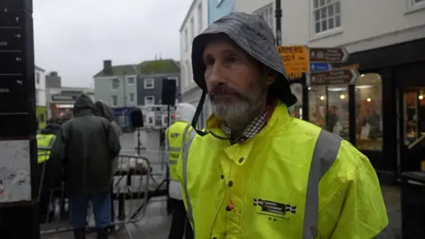 Peter Lawrence pictured in a high-vis jacket in front of the protest. Behind him people are standing, and there are signs on a signpost.