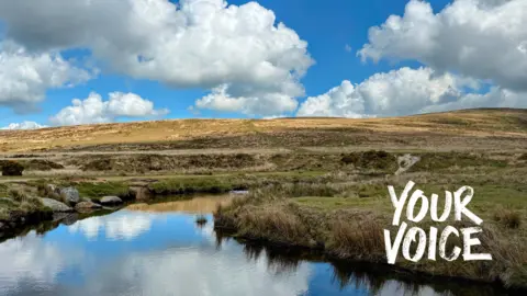 A stream near Belstone on Dartmoor with hills in the distance and a cloudy sky with the Your Voice branding overlaid on the lower right corner of the image