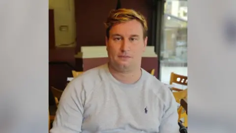 Peter Lawriw Bahan Alexander Paul Burton wearing a grey top and looking at the camera. 