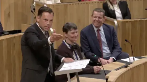 A man in a black suit is gesturing towards someone off screen in the Senedd, while a woman in a brown cardigan and a man in a blue suit with red tie are in the background smiling. There are also wooden seating areas and wired microphones coming out of the in built wooden desks within the Senedd. 