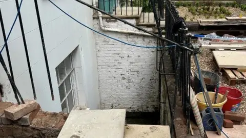 An image of a basement lightwell seen from street level, with railings missing on the right hand side of stone steps. The terraced property's walls are painted white.