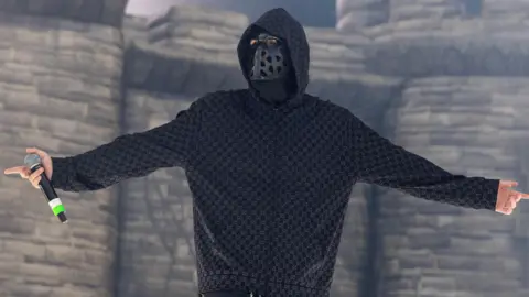 EsDeeKid performs in front of a blow-up castle during a set in Illinois.  He is seen looking at the crowds while holding a microphone and wearing a black mask with holes and a hoodie.