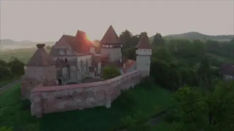 Mihai Eminescu Trust A castle and walls sitting on a green hill, surrounded by green space. The sun is rising in the background