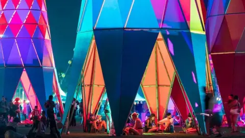 Iwan Baan Sarbalé Ke structures with hollow interior and colourful patterns on them. There are people sitting inside the hollow interior.