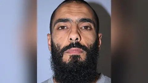 Greater Manchester Police Police custody image of Bilel Saadaoui. He has short black hair and a long black beard and is wearing a grey top.
