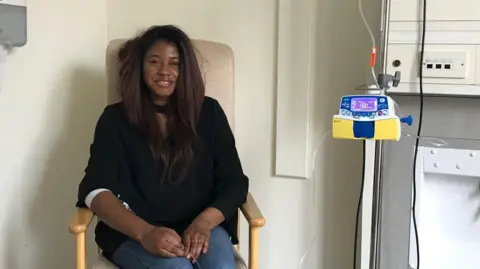 Norfolk and Norwich University Hospital A woman sits in a chair in a hospital room, while receiving treatment. She wears a black top and blue denim jeans. She appears to be hooked up to an intravenous - a white tube extends down to her finger from a yellow machine. 