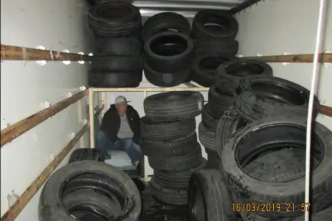 Home Office A wider view of the vehicle's interior shows one man sitting inside a makeshift compartment among numerous stacks of car tyres. The wooden panels of the compartment are clearly visible, and the space appears dimly lit. One man’s face is blurred for anonymity. The timestamp "16/03/2019 21:57" is visible in the bottom corner of the images.