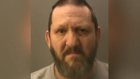 Police mugshot of a man with short brown hair and greying brown beard. He has dark mole under one of his eyes. 