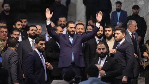 Nabieha Altaha/Anadolu via Getty Image President Ahmed al-Sharaa, wearing a navy suit and blue tie, raising his hands in the air, at an event in November 2025 to celebrate the anniversary of the end of the Assad regime's control of Aleppo. He has dark hair and a dark beard, and is surrounded by other men in suits.
