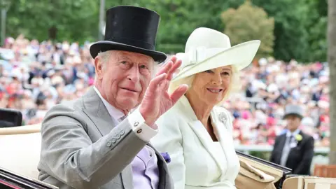 King Charles wearing a grey suit and black top hat and Queen Camilla in a cream-coloured outfit smiling and waving. 