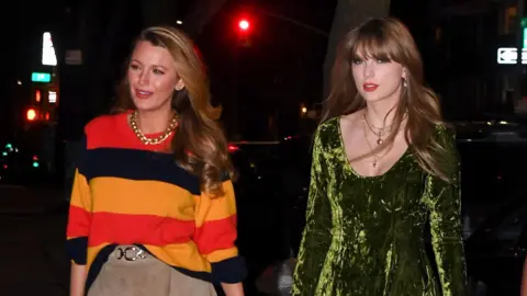Blake Lively, wearing an orange, red and black stripy jumper and a tan suede skirt, walking alongside Taylor Swift, who is in a green crushed velvet dress