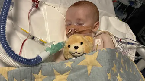 Family Photo Baby asleep in hospital bed surrounded by tubes and wires - and a teddy bear
