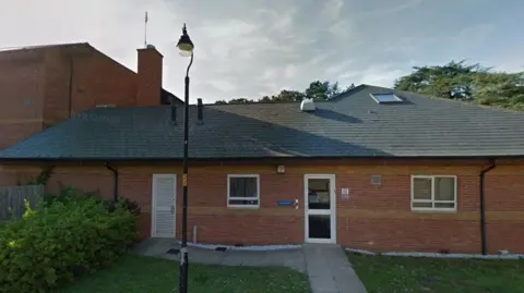 Google Brick-built single-storey hospice building with pitched grey roof. A two-storey building is visible to the left and there are trees to the right. There is a grass lawn in front of the hospice.