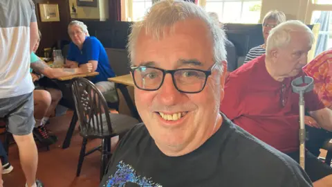 BBC/ Carla Fowler A man with glasses wearing a black t shirt smiles from inside a pub.