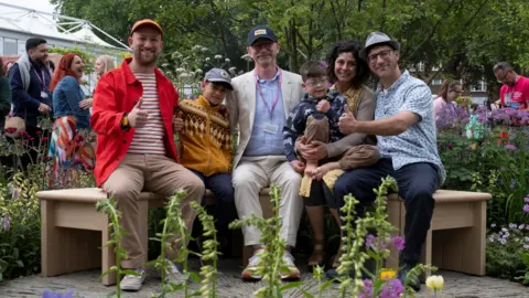 Ginger Horticulture A family sit with the designers of the garden on a bench surrounded by flowers and plants at the Chelsea flower show.