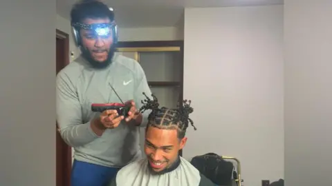 A man wearing a head torch trims another man's hair with clippers in a room with a wooden cupboard and metal-framed chair visible in the background.