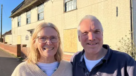 Debbie, on the left, and Mike on the right. She is wearing a beige pullover, and white teeshirt beneath. She wears glasses, and has long shoulder-length blonde hair. Mike has greying hair, slightly receding, and is wearing a blue polo shirt and white teeshirt beneath. Both are smiling, standing in front of the fire-damaged Gables Farm, which they have purchased.