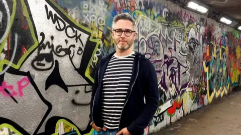 SDLP Councillor Gary McKeown stands in front of wall covered in graffiti