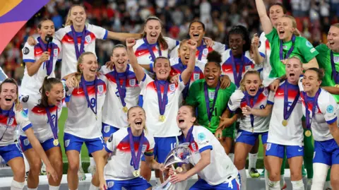 England's Leah Williamson and Keira Walsh lift the trophy as they celebrate with teammates after winning the UEFA Women's Euro 2025. All the players are in their white kit with blue shorts and have medals around their necks. They are cheering and smiling.