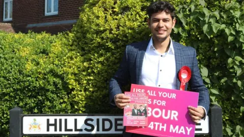 Tushar Kumar A neatly dressed person, Tushar Kumar, stands on a quiet residential street, framed by lush green hedges. The sky above is bright and clear blue, suggesting a calm, sunny day. Behind Kumar, a street sign reads “HILLSIDE AVENUE,” Kumar is holding a bold pink campaign sign with white lettering that reads “USE ALL YOUR VOTES FOR LABOUR 4TH MAY,” along with a leaflet featuring smaller images and additional names and text. 