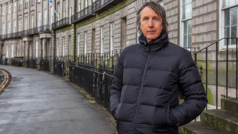 HOLYROOD PR Philip Flockhart has long hair and is wearing a blue puffer jacket. He is standing on front of the crescent of Georgian houses.