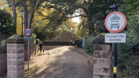 A winding narrow road with a sign at the entrance saying no entry to cars and motorbikes except for access and trees lining both sides