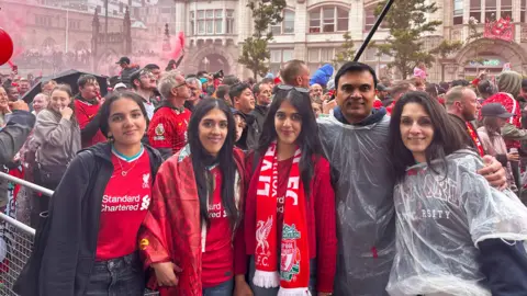 Harry Rashid A man and woman stand with three younger women, in front of a crowd on a street. The three younger women are wearing red football shirts. Many people in the crowd behind the are also wearing red football shirts.