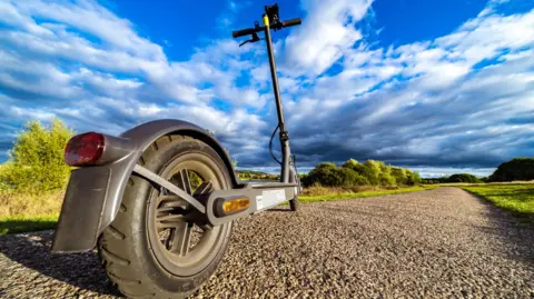 CHUNYIP WONG/Getty Images A close-up of an e-scooter on a sunny day. The vehicle is black with two black wheels, a narrow black steering column, a red rear light and an amber side light on the wheel. A gravel path lined by grass and trees stretches into the distance in front of the e-scooter. 