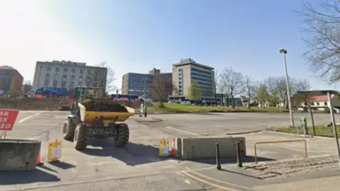 Google A construction vehicle with a trailer full of compost sits on an empty car park. Cones, bollards and other street furniture is visible.