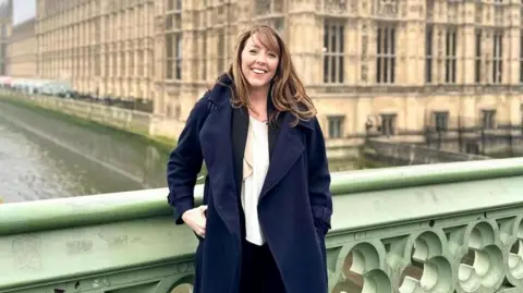 Katie Rollings A woman with brown hair wearing a white shirt under a navy trench coat. She is standing in front of the Houses of Parliament. 