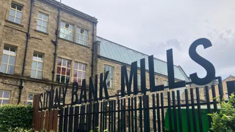 BBC/ Elizabeth Baines A yellow brick building in the background, with a small hedge in front. There is a sign saying "Sunny Bank Mills" on top of a black metal fence.