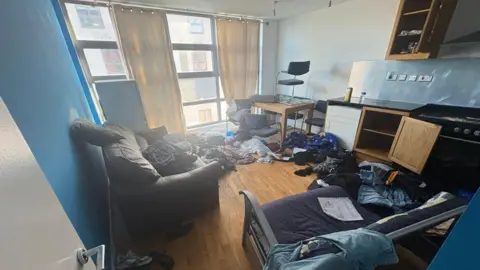 Contributed An open plan living and kitchen area. The walls are painted blue and white. There are items like clothing discarded across the floor and sofas. Some of the cupboard doors have been left open in the kitchen area.