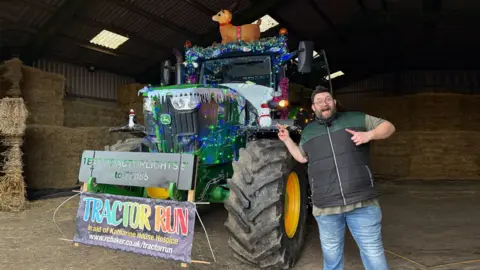 BBC Radio Oxford presenter Adam Ball stands in front of a christmas decorated tractor in a barn.
