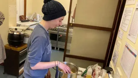 Dave Webster/BBC Aimee Clasby, trainee youth development worker at a baking session. She wears a blue sweatshirt and a black bobble hat and is mixing some food in a saucepan.