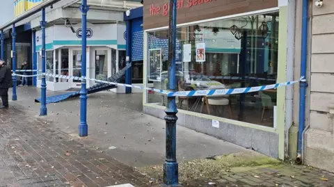 Eddie Mitchell A row of shops is cordoned off, with metal shutters which have been smashed off.
