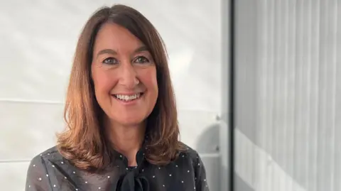 Alison is seated indoors on a black office chair, wearing a sheer black blouse with white polka dots and a bow tie detail at the collar. 
