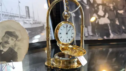 Hannah Roe/BBC A gold pocket watch on display. There are several black and white photos behind it.