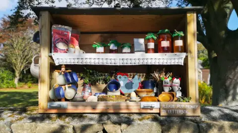 A wooden 'hedge veg' stall which has jars of preserves and pottery mugs for sale. It is sitting on a wall with trees in the background.