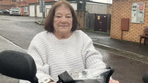 Nickie Smith is on a mobility scooter looking at the camera with a stern face. She is on a residential road that is wet with rain.