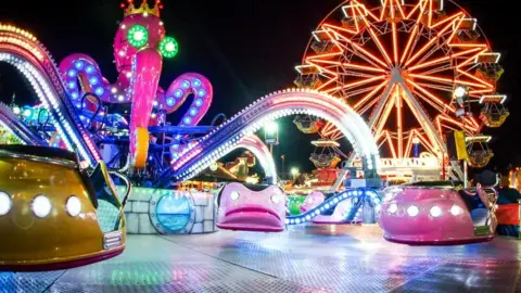 A selection of brightly-coloured fairground rides are pictured, with a ferris wheel in the background