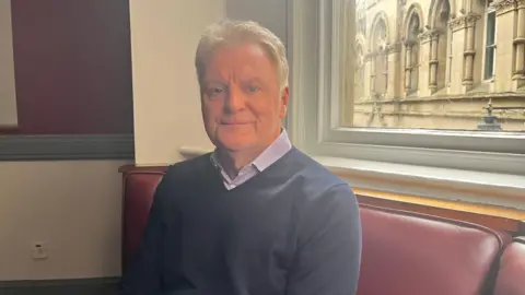 Steven Howard sat on a sofa with an old building outside behind him