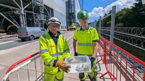 RSPCA Two men in yellow reflective clothing and safety helmets with a translucent plastic box with two birds inside and coloured guard rails on both sides of them and a large industrial building behind them