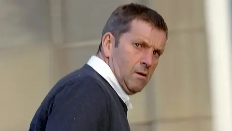Close-up of a person in a dark sweater turning their head to look over their shoulder, with a blurred outdoor background