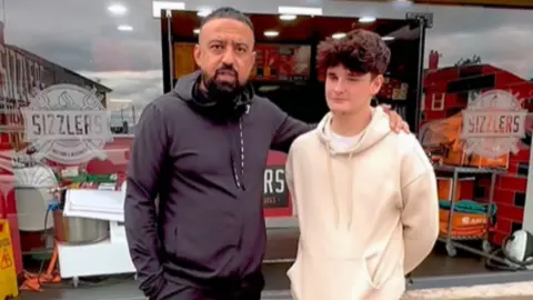 Arfan and Tyler stand together outside Sizzlers.  Arfan has his arm around Tyler. Tyler has short brown hair and and Arfan is wearing a black top.