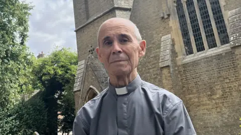 John Devine/BBC Richard Hines is standing in front of a stone church. He is wearing clerical clothing and has short hair.