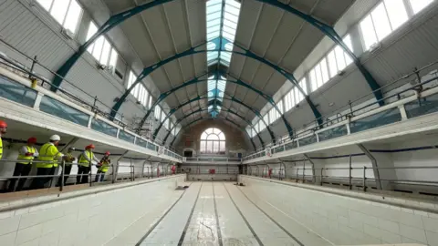 Swindon Borough Council A rectangular room with a large swimming pool down the centre. The pool is deep with white tiles which are cracked on the floor. Along the pool floor, there are 5 black lines signalling the swimming lanes. Above the pool is a big curved, Victorian window made of stain glass. On all sides of the pool are scaffolding for the continuing work.